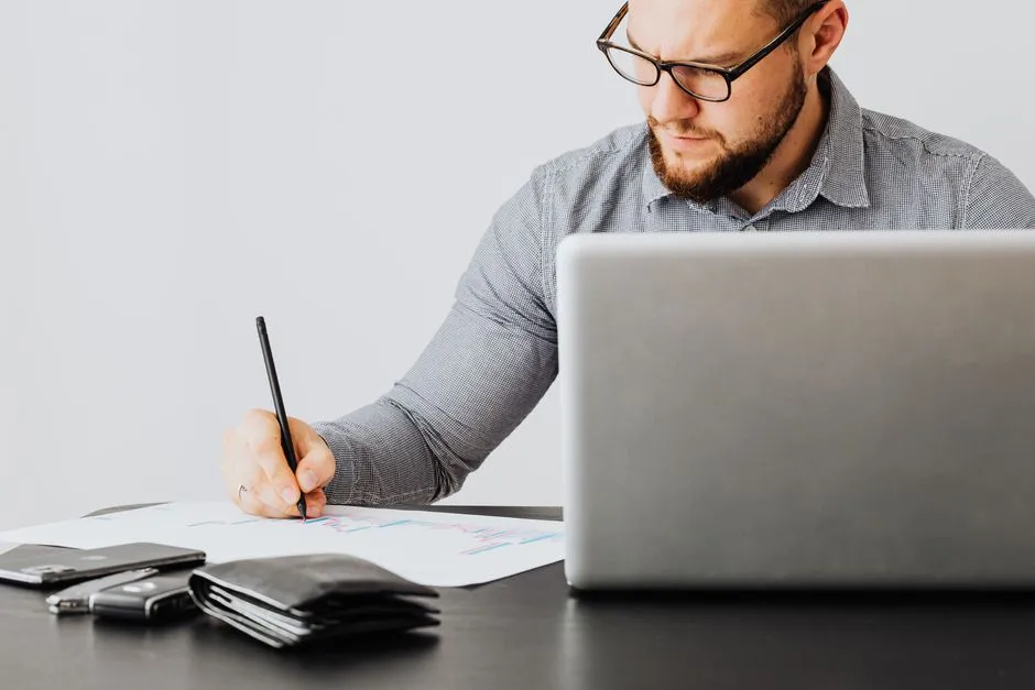 A focused man analyzing financial papers with a laptop in an office setting.